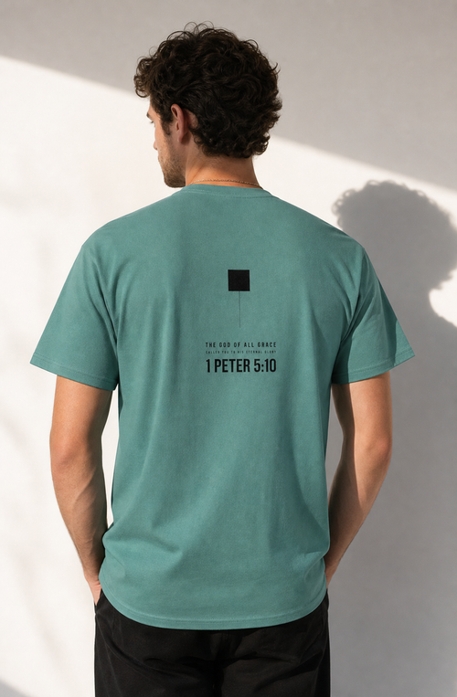 A man viewed from behind wearing a teal-green t-shirt with a black graphic print on the back reading 'I PETER 5:10' and 'ONE KINGDOM OF ALL SCRIPTURES'.