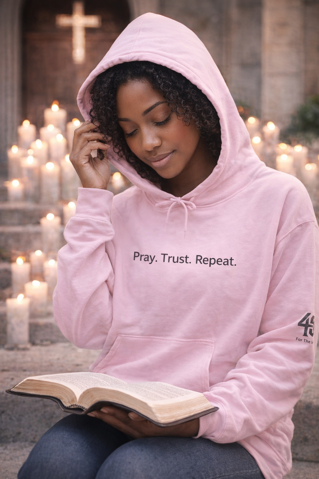 Person wearing a pink hoodie with 'Pray. Trust. Repeat.' text, holding a book outdoors with candles in the background.