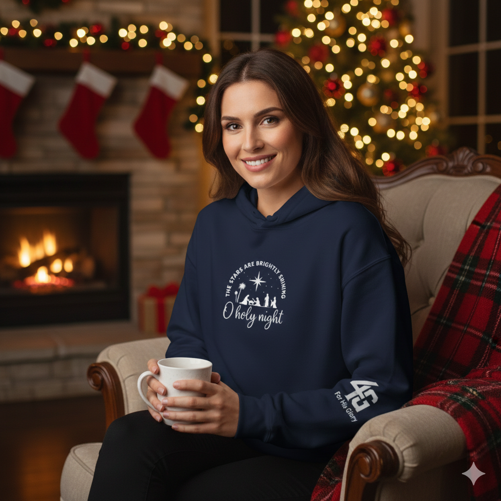 Woman in a navy blue hoodie with a Christmas-themed design, holding a mug, sitting in a cozy room with a fireplace and decorated Christmas tree.