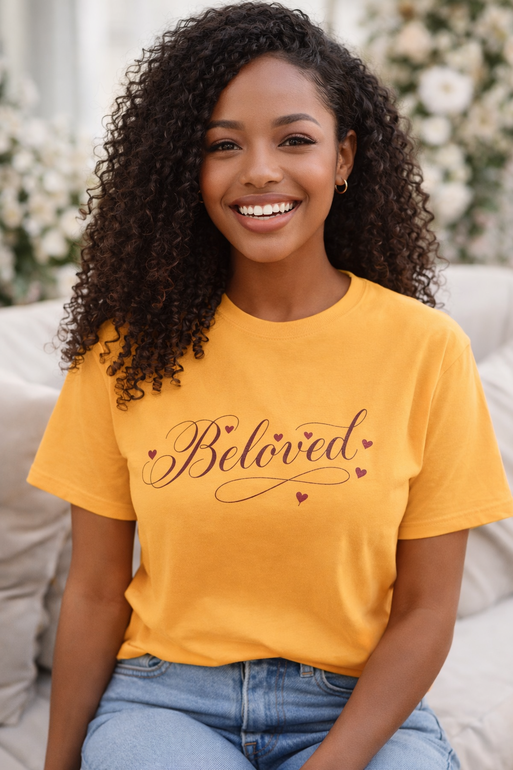 Woman wearing a yellow t-shirt with 'Beloved' printed on it, sitting in a decorated room.