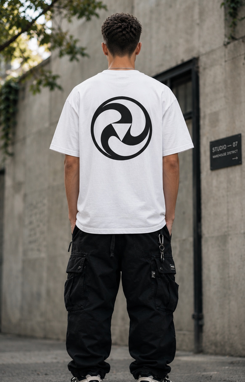 A man viewed from behind wearing a white t-shirt with a large black circular graphic on the back, paired with black cargo pants, standing outdoors against a concrete wall.
