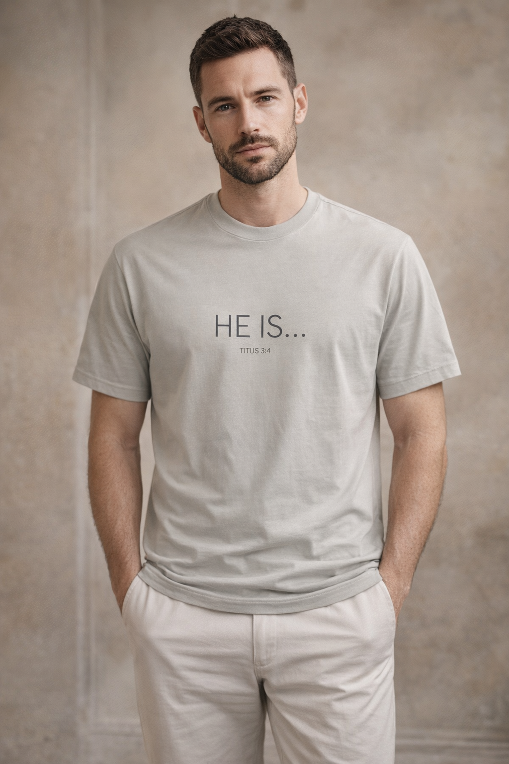 A man wearing a light gray crew-neck t-shirt with the text 'HE IS.