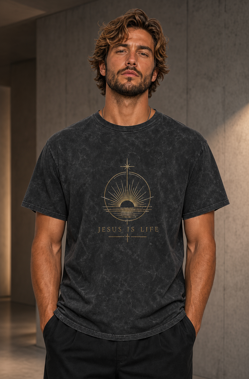 A man wearing a dark gray mineral-wash t-shirt with a gold graphic of a rising sun and the text 'JESUS IS LIFE' on the front, standing in a modern interior hallway.