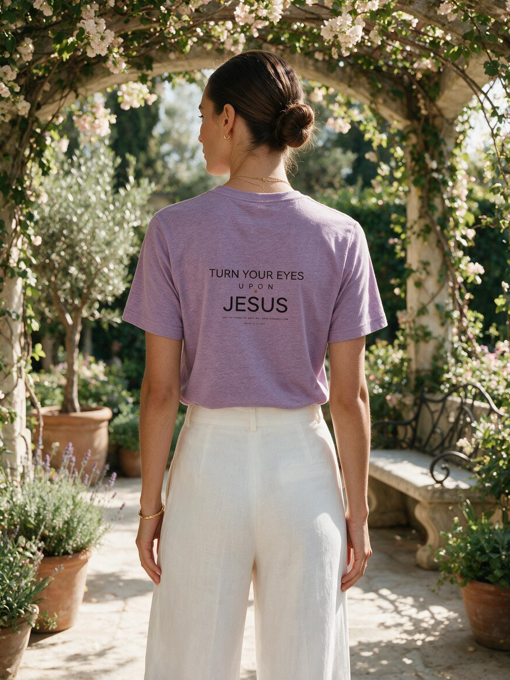 A woman viewed from behind wearing a lavender graphic t-shirt with the text 'TURN YOUR EYES UP JESUS' printed on the back, paired with white wide-leg pants, standing in a sunlit garden under a flowering arbor.