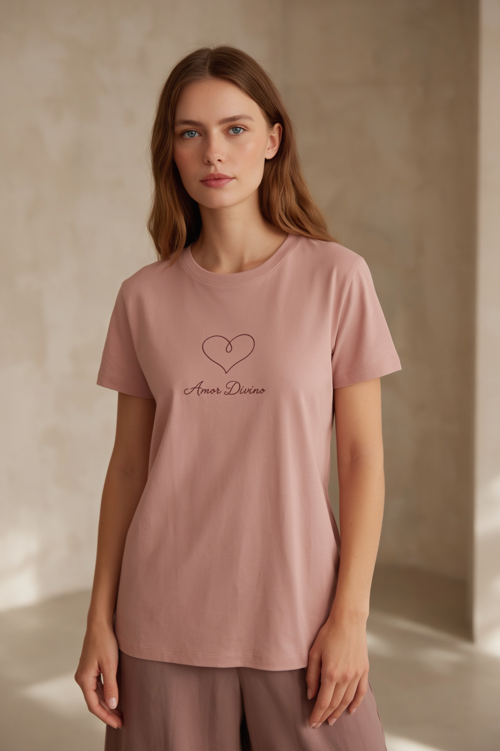 A woman wearing a dusty pink short-sleeve t-shirt with a black heart outline and the words 'Amor Divino' printed on the chest, paired with matching dusty pink trousers, set against a warm beige textured wall.