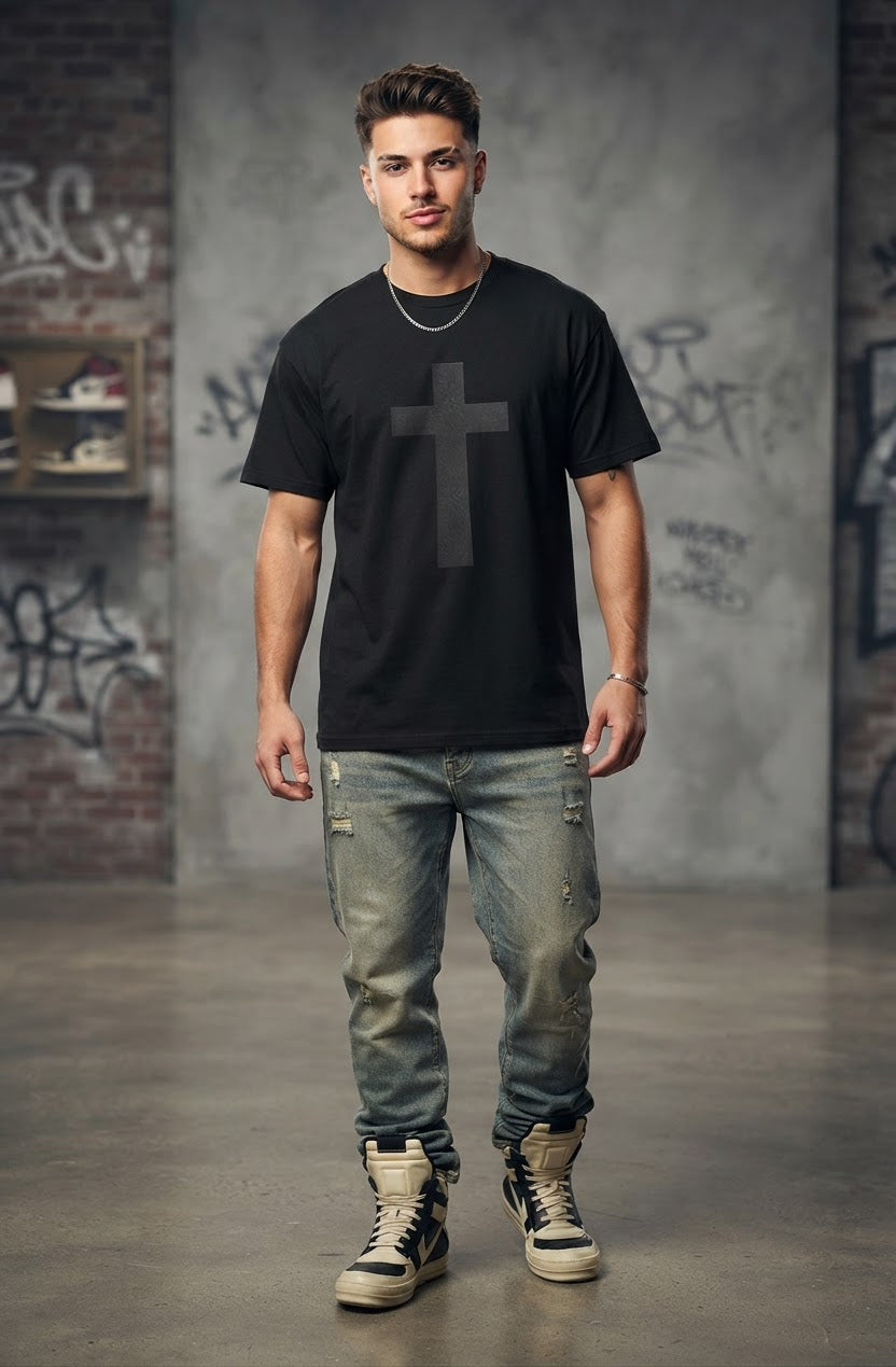 A man modeling a black crewneck t-shirt with a large dark gray cross graphic on the front, paired with distressed blue jeans and beige high-top sneakers, standing in a concrete warehouse with graffiti walls.
