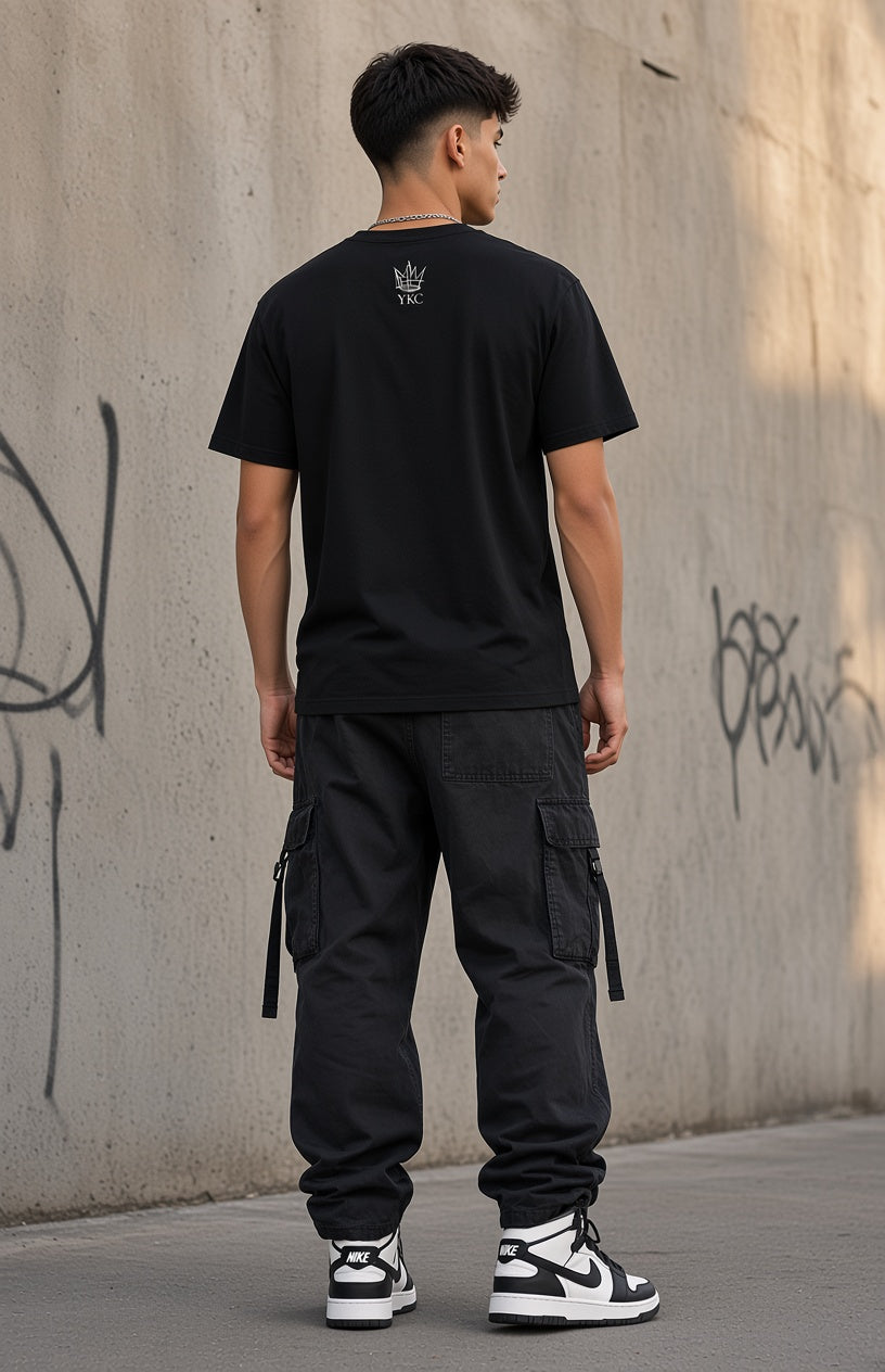 A man viewed from behind wearing a black crewneck t-shirt with a small white graphic on the back, black cargo pants, and black and white sneakers, standing against a concrete wall with graffiti.