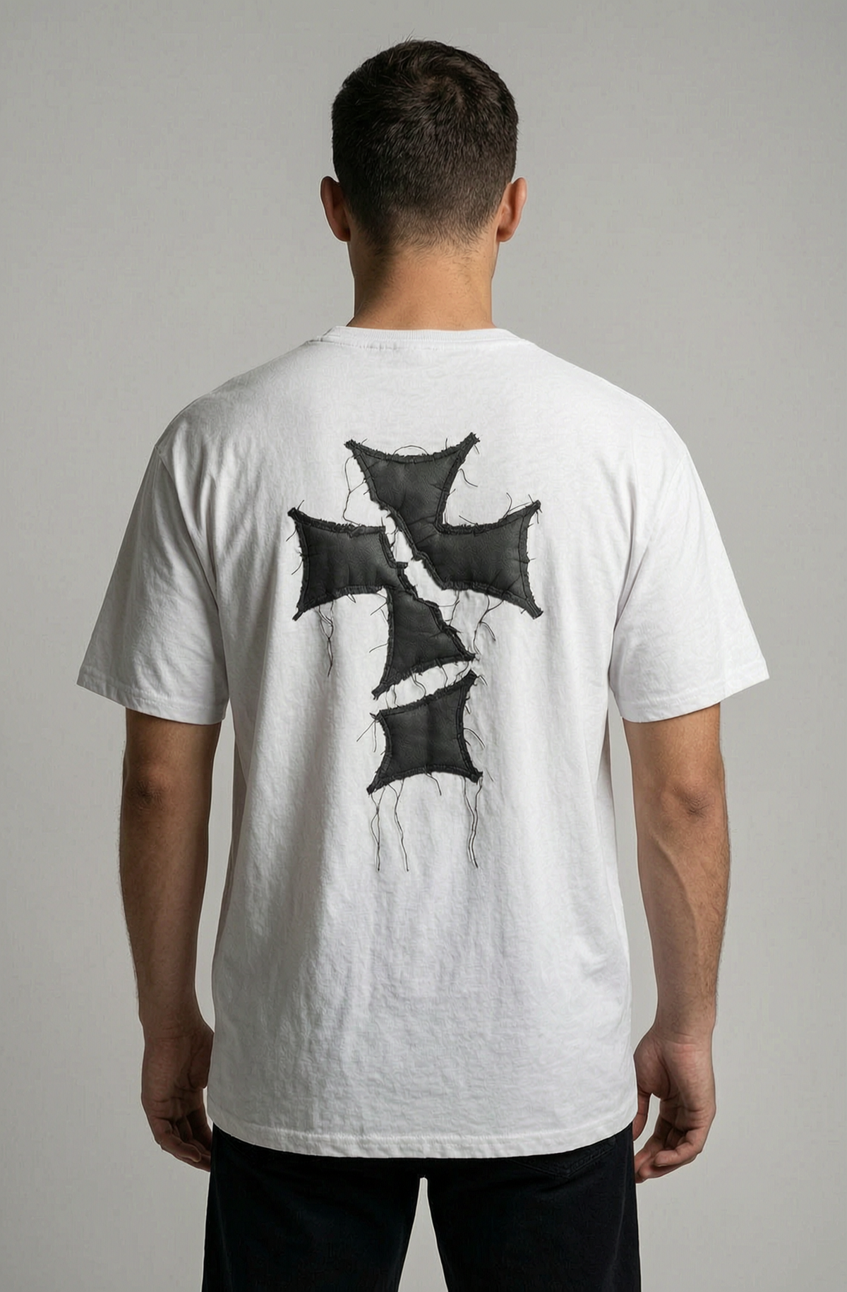 A man viewed from behind wearing a white t-shirt with a large black distressed cross graphic on the back, paired with black pants, against a neutral gray background.