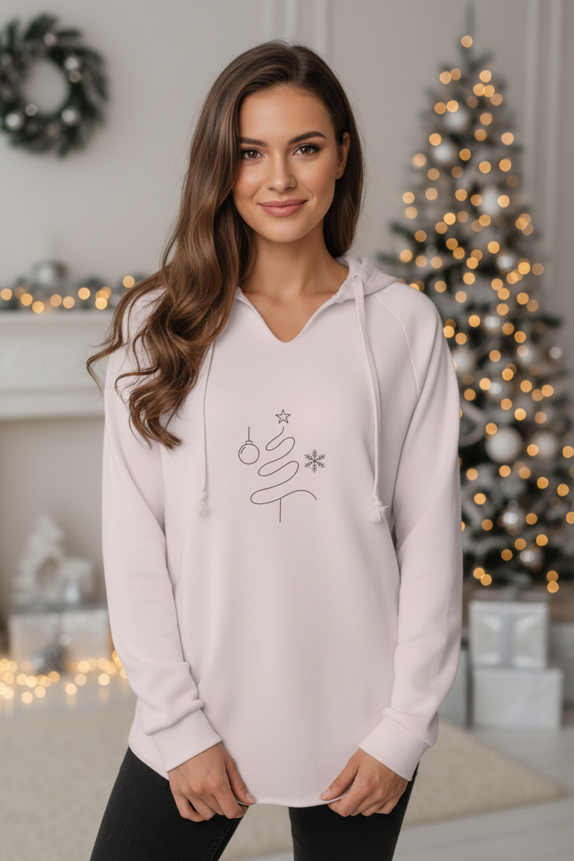 A woman wearing a light pink pullover hoodie with a simple black line-art Christmas tree graphic on the front, posed in a cozy, softly lit holiday home setting with a decorated Christmas tree in the background.