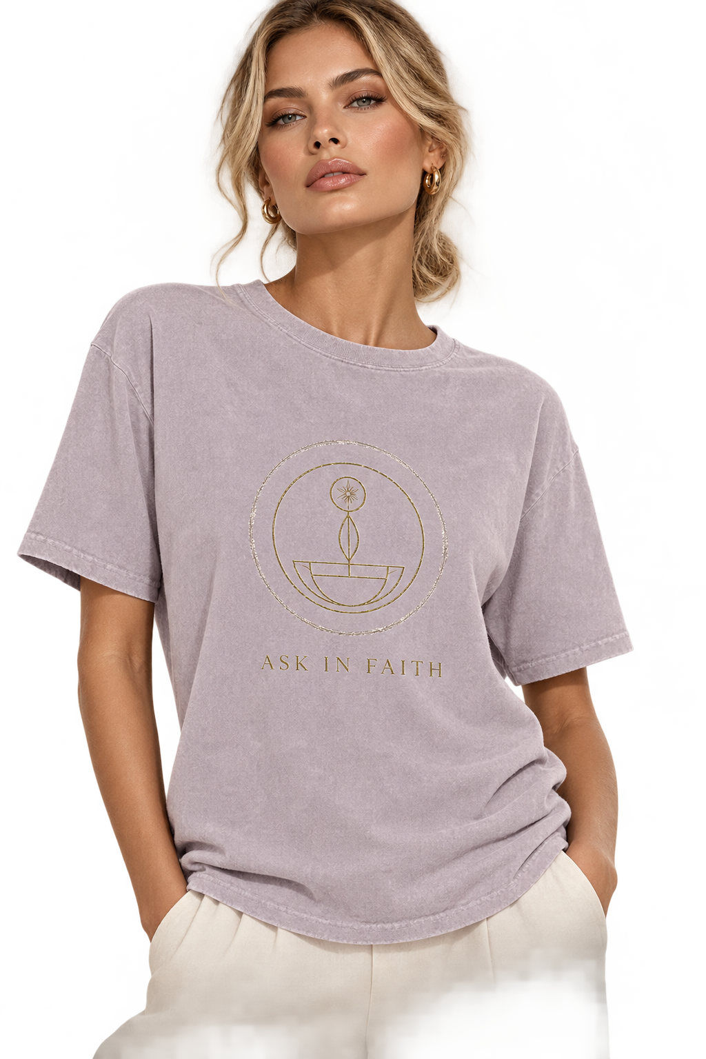 A woman wearing a light lavender-gray short-sleeve t-shirt with a gold circular graphic and the words 'ASK IN FAITH' printed on the front, paired with off-white pants against a white studio background.