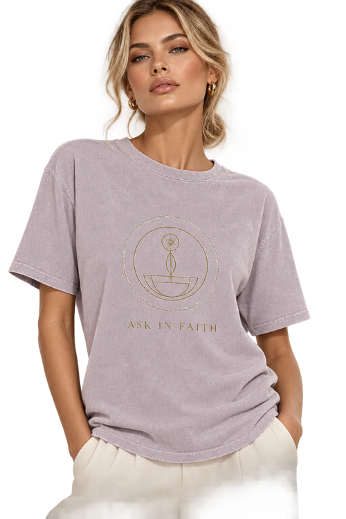 A woman wearing a light lavender-gray short-sleeve t-shirt with a gold circular graphic and the words 'ASK IN FAITH' printed on the front, paired with off-white pants against a white studio background.