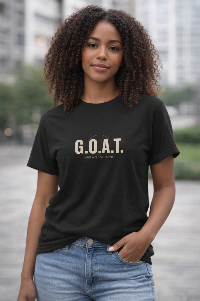 A woman with curly brown hair wearing a black short-sleeve t-shirt with 'G.O.A.T.