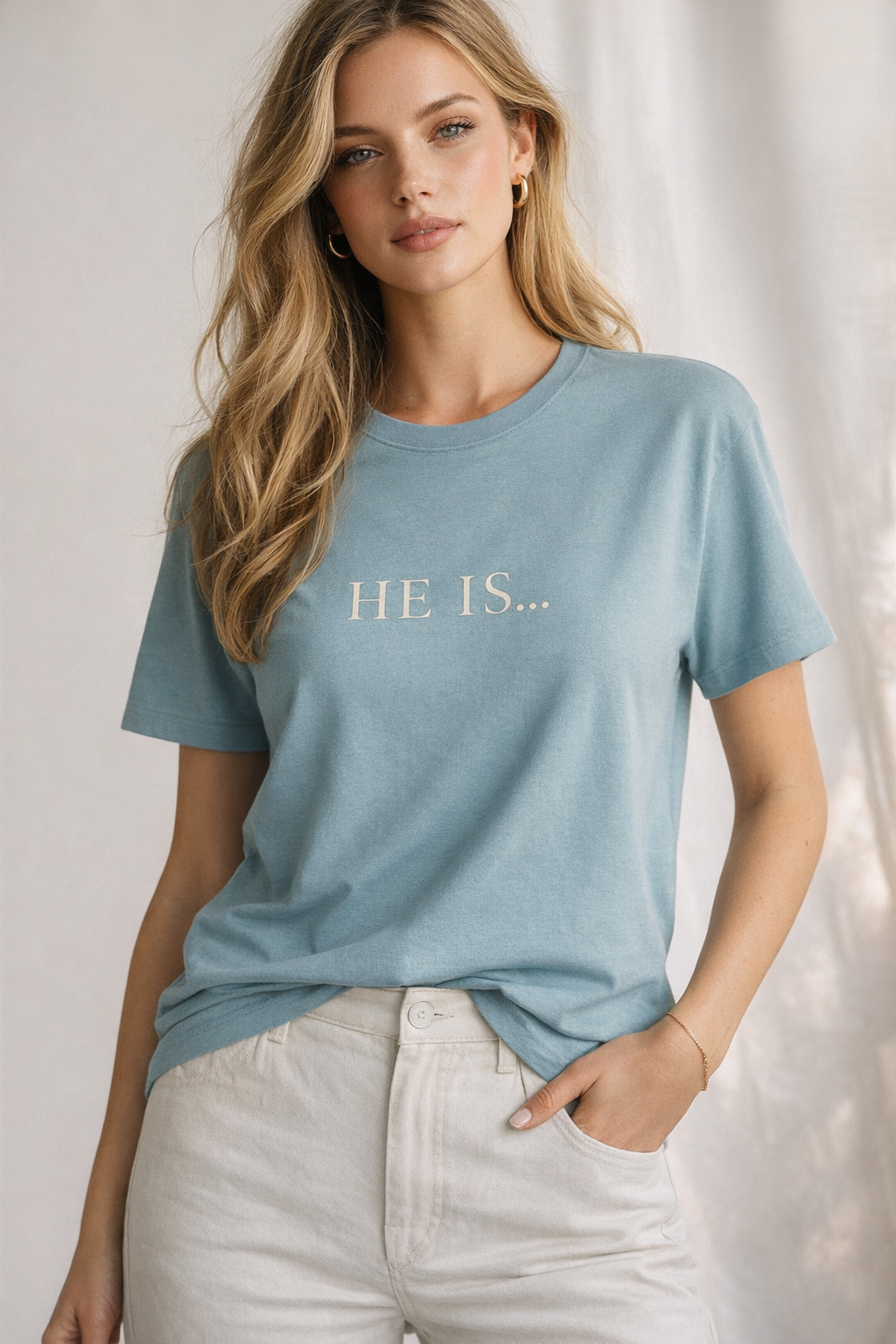 A woman wearing a light blue crewneck t-shirt with 'HE IS.