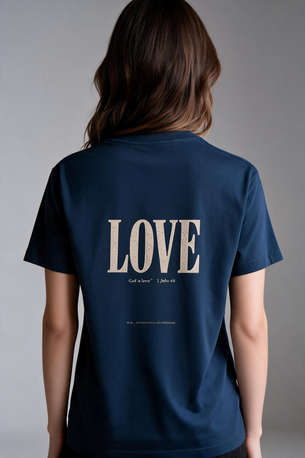 A woman viewed from behind wearing a navy blue t-shirt with the word 'LOVE' printed in large beige serif letters on the back, with smaller scripture text underneath.
