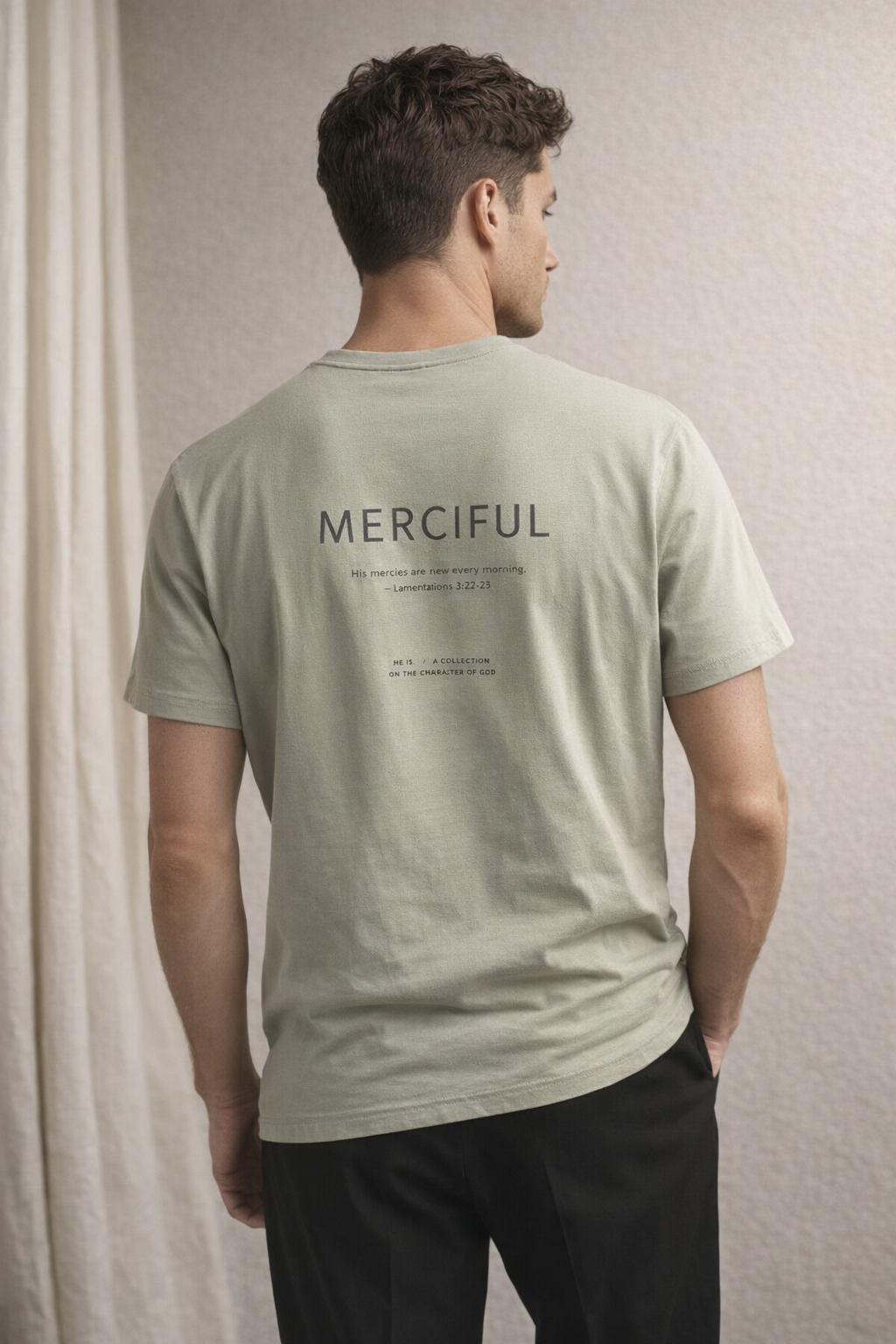 A man shown from behind wearing a light sage green t-shirt with 'MERCIFUL' printed in black text on the back, paired with black pants.