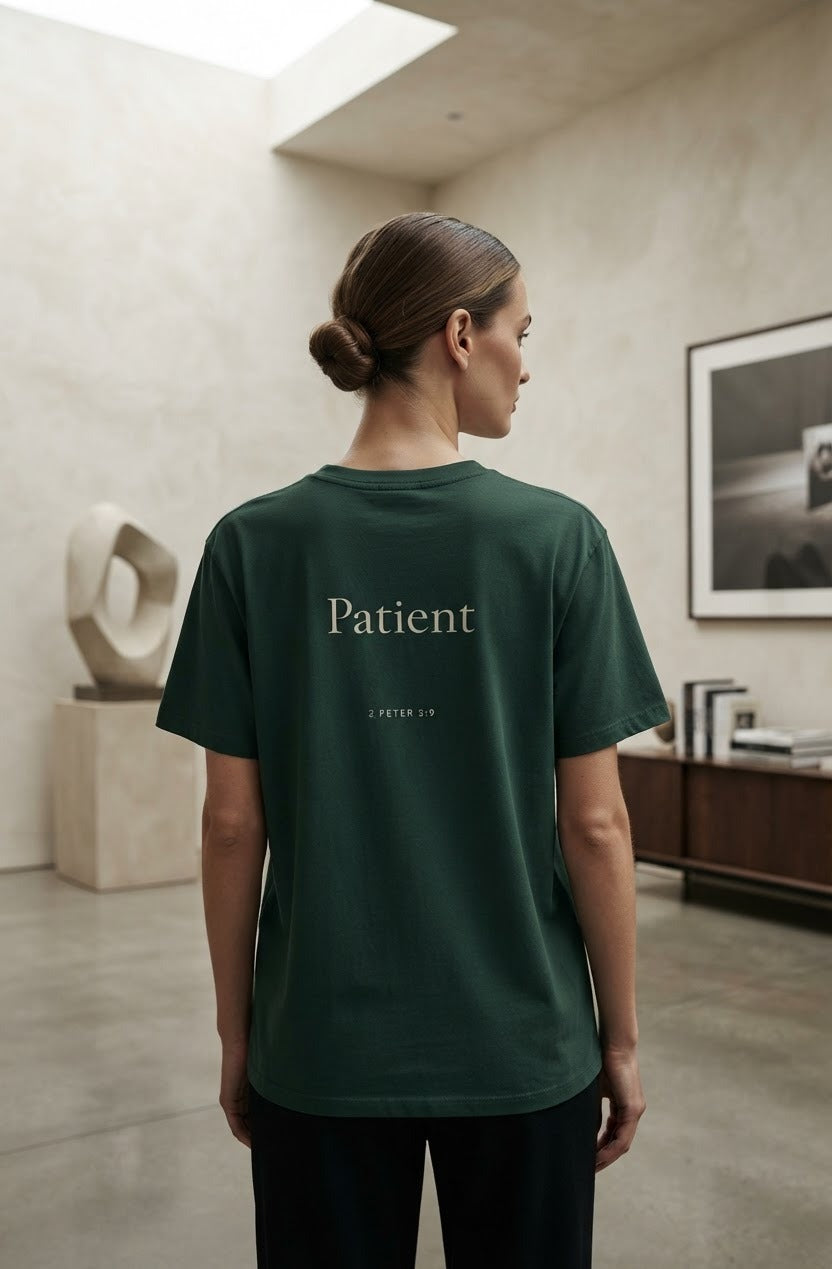 A woman viewed from behind wearing a dark forest green t-shirt with the word 'Patient' printed in white serif font on the back, standing in a minimalist, modern interior room.