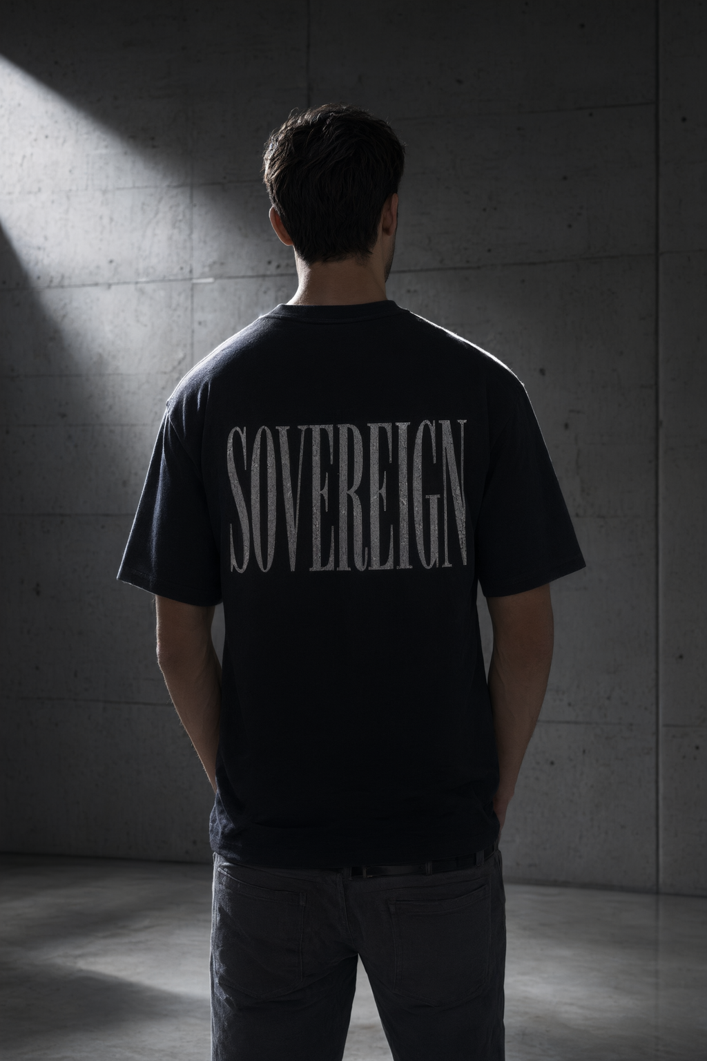 A man viewed from behind wearing a black t-shirt with 'SOVEREIGN' printed in a white serif font across the back, standing in a dimly lit concrete room.