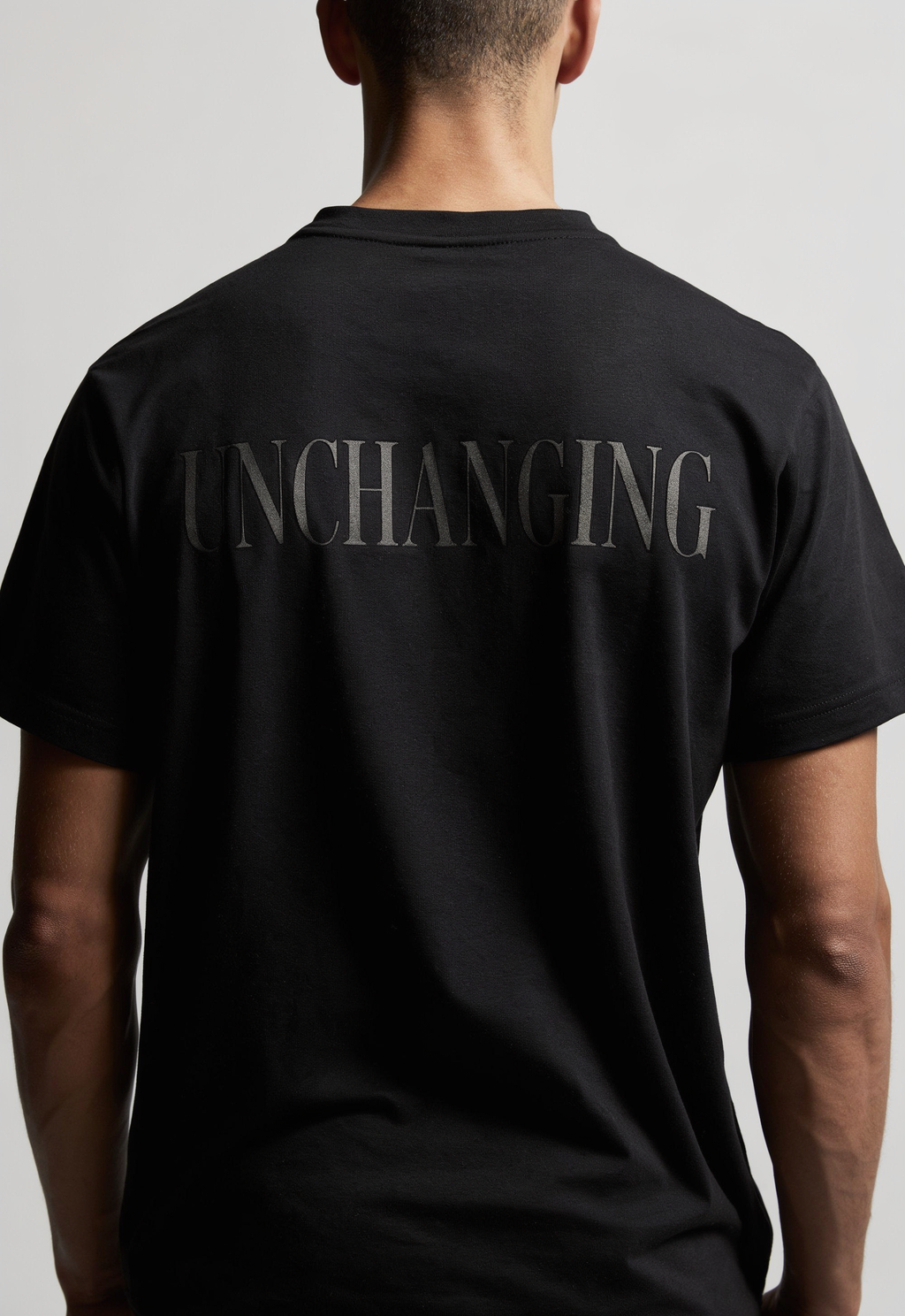 A man viewed from the back wearing a black short-sleeve t-shirt with the word 'UNCHANGING' printed across the upper back in a subtle dark gray tone.