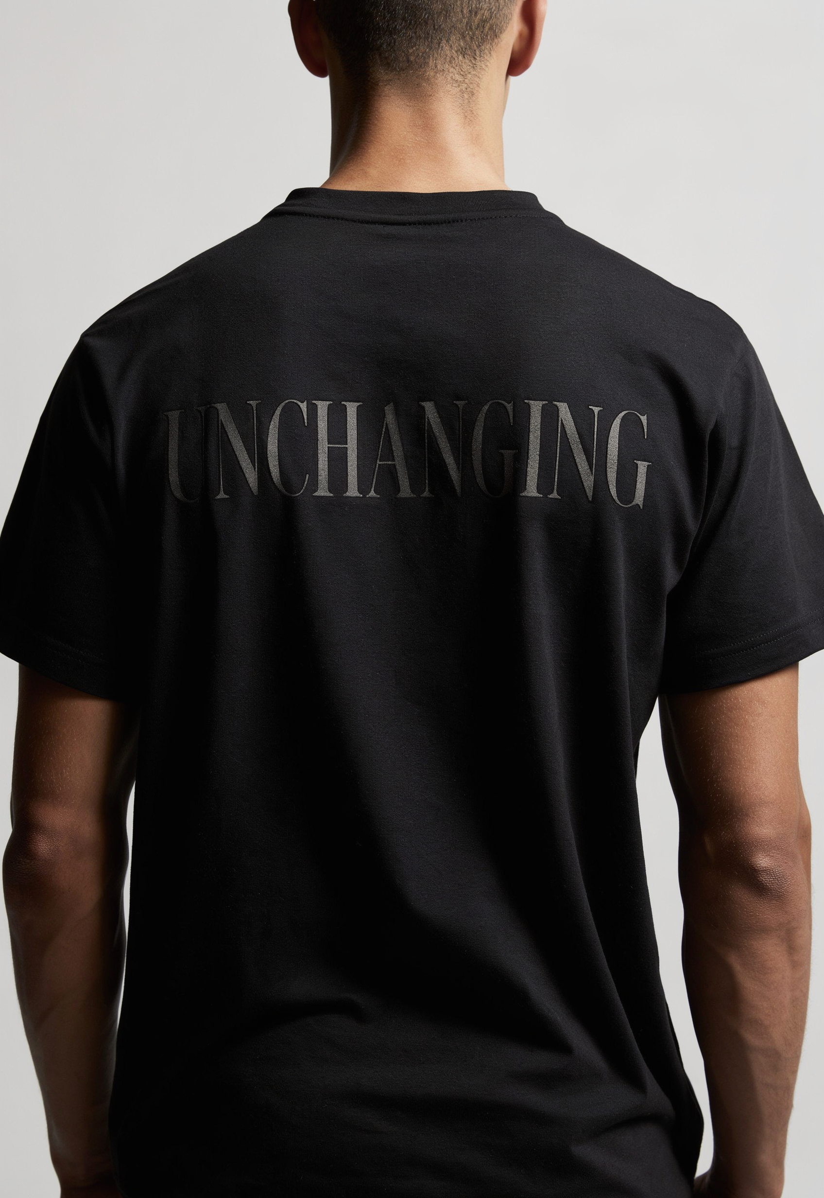 A man viewed from the back wearing a black short-sleeve t-shirt with the word 'UNCHANGING' printed across the upper back in a subtle dark gray tone.