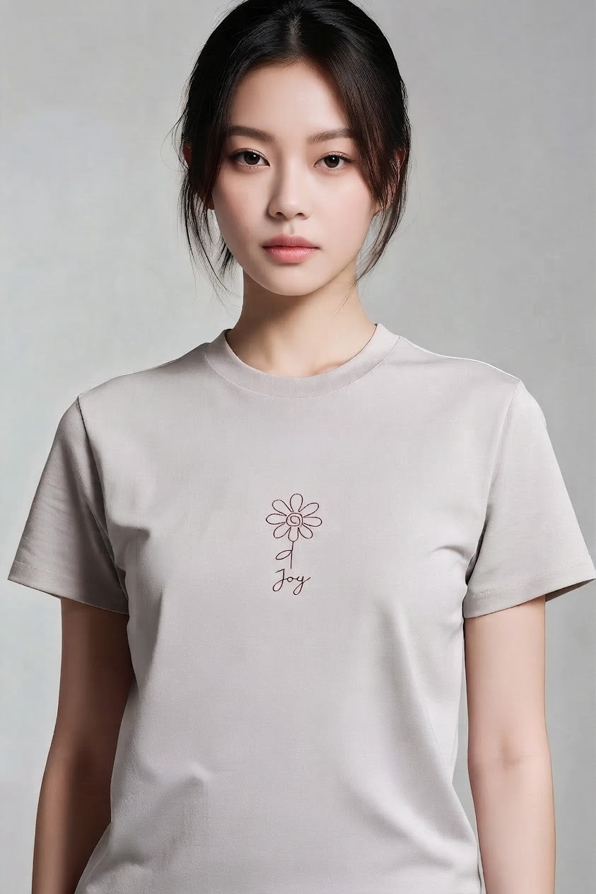 A woman wearing a light gray crewneck t-shirt with a small pink floral graphic and the word 'Joy' printed on the chest, set against a neutral gray background.