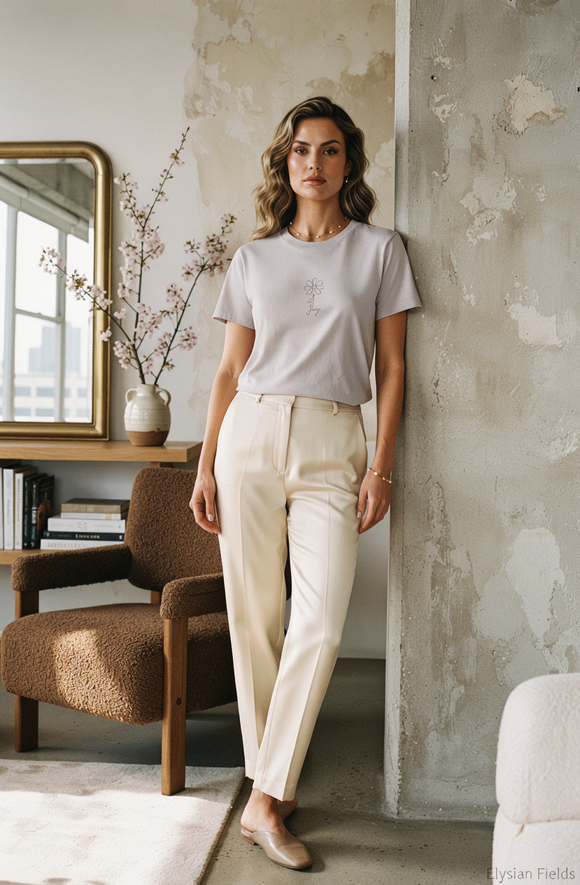 A woman stands indoors wearing a light gray short-sleeve t-shirt and cream-colored straight-leg trousers, posed next to a textured concrete wall in a modern loft setting.