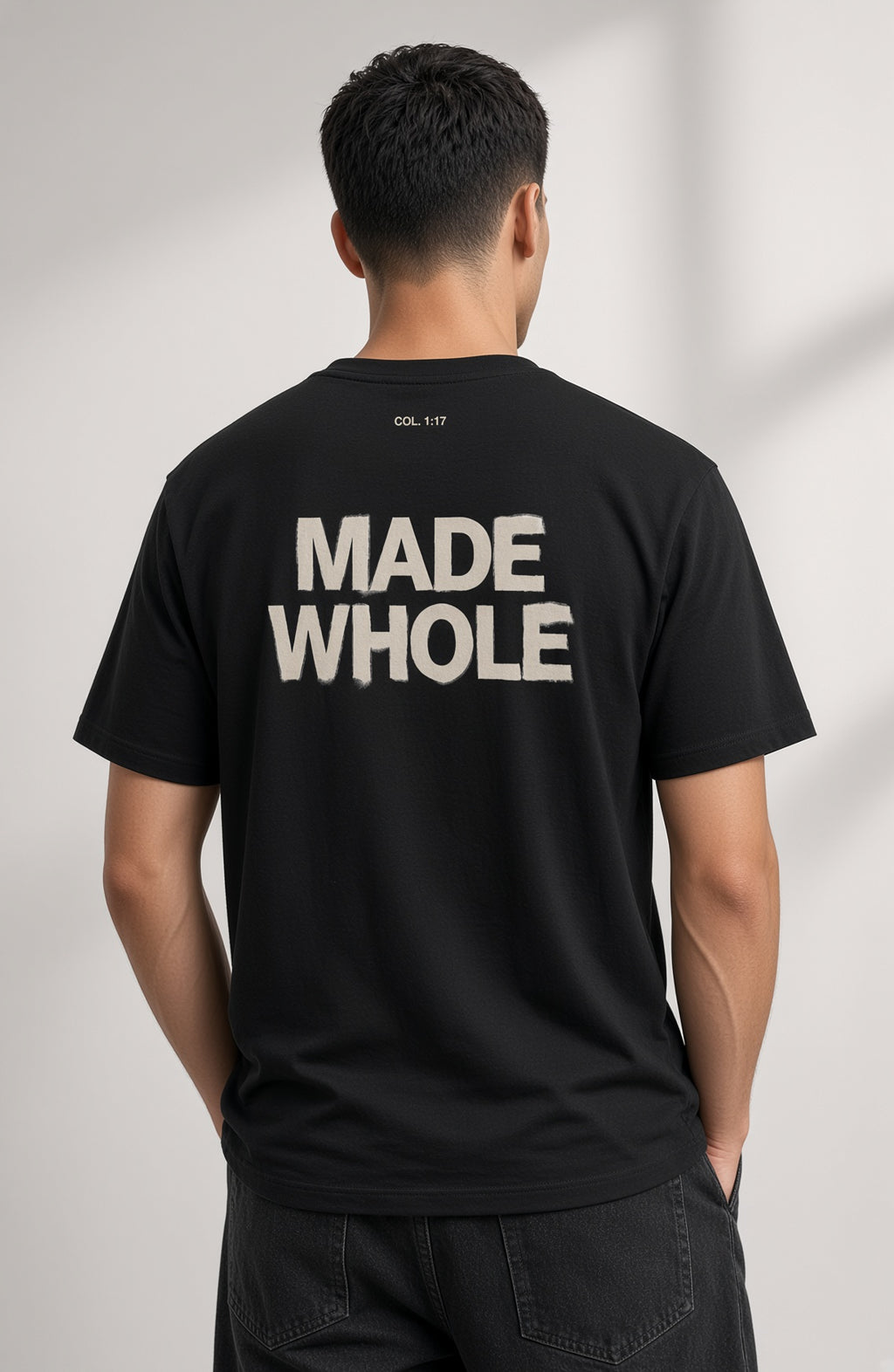 A man viewed from behind wearing a black t-shirt with 'MADE WHOLE' printed in large white letters on the back, paired with dark jeans against a light gray background.