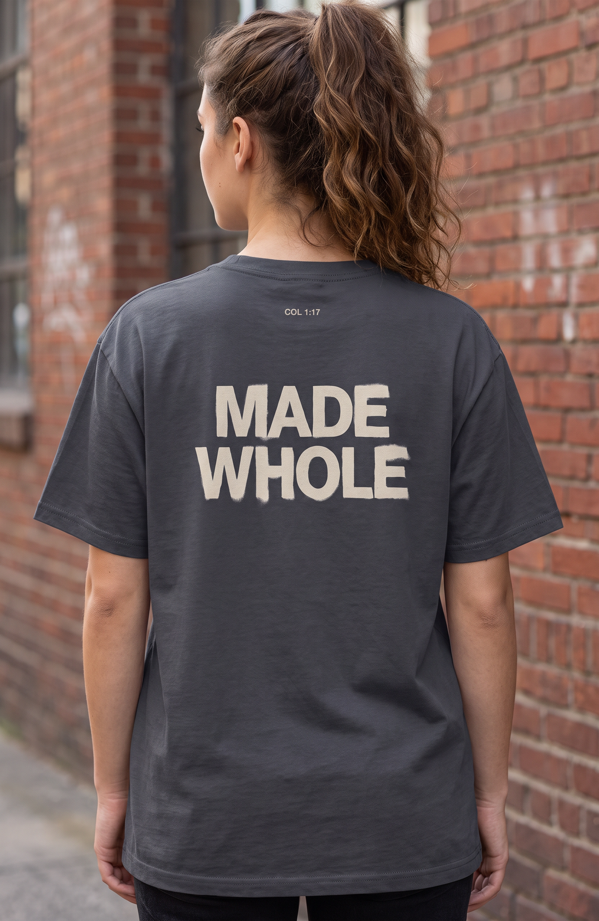 A woman viewed from behind wearing a dark charcoal gray t-shirt with 'MADE WHOLE' printed in bold cream letters on the back, standing against a red brick wall.