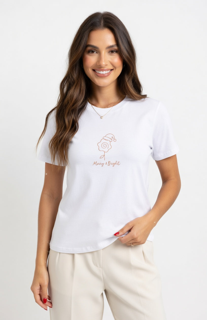 A woman modeling a white short-sleeve crewneck t-shirt with a small rose gold line-art Christmas ornament graphic and the text 'Merry & Bright' on the chest, paired with beige pants against a plain white studio background.