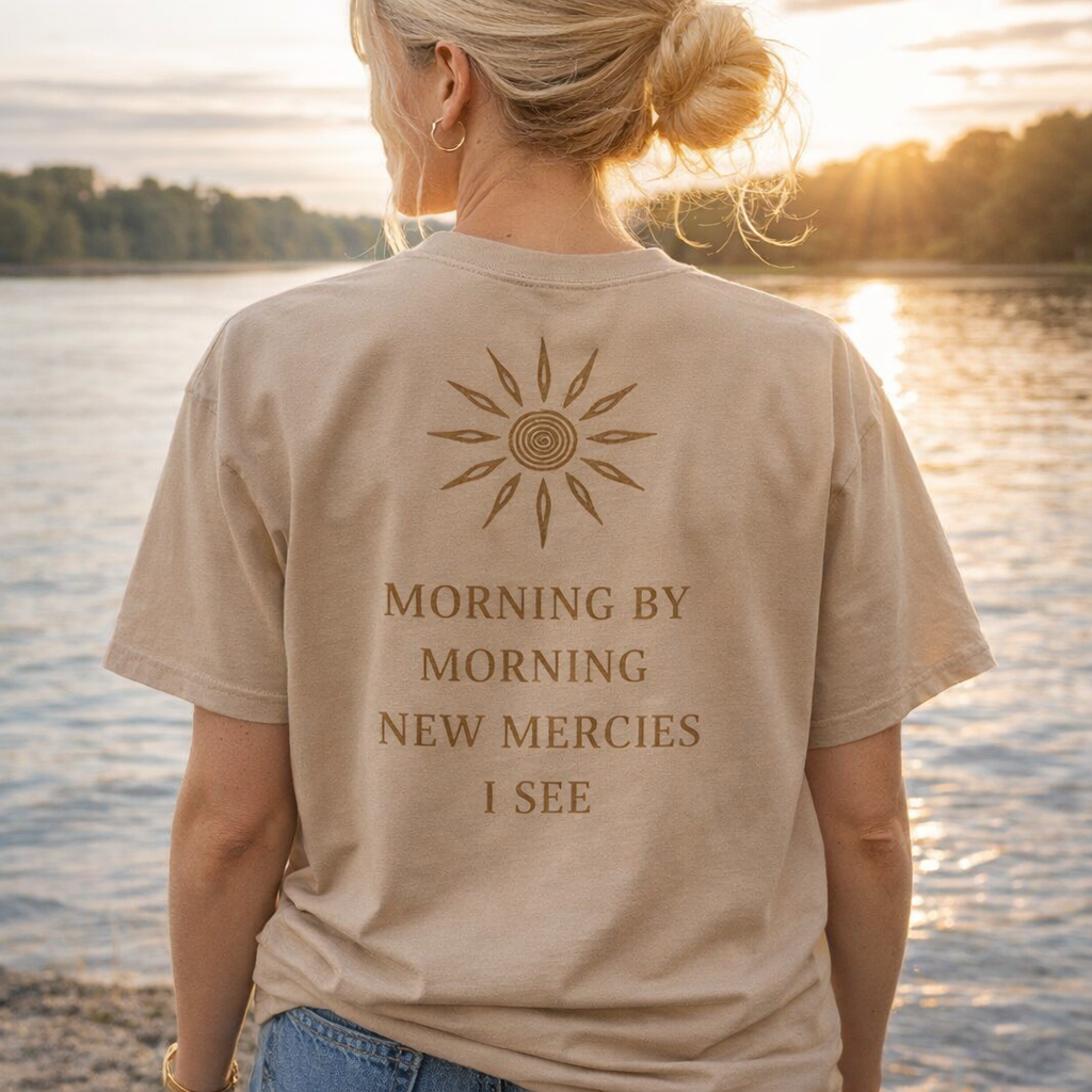 Person wearing a beige t-shirt with a sun design and text by a lake at sunset