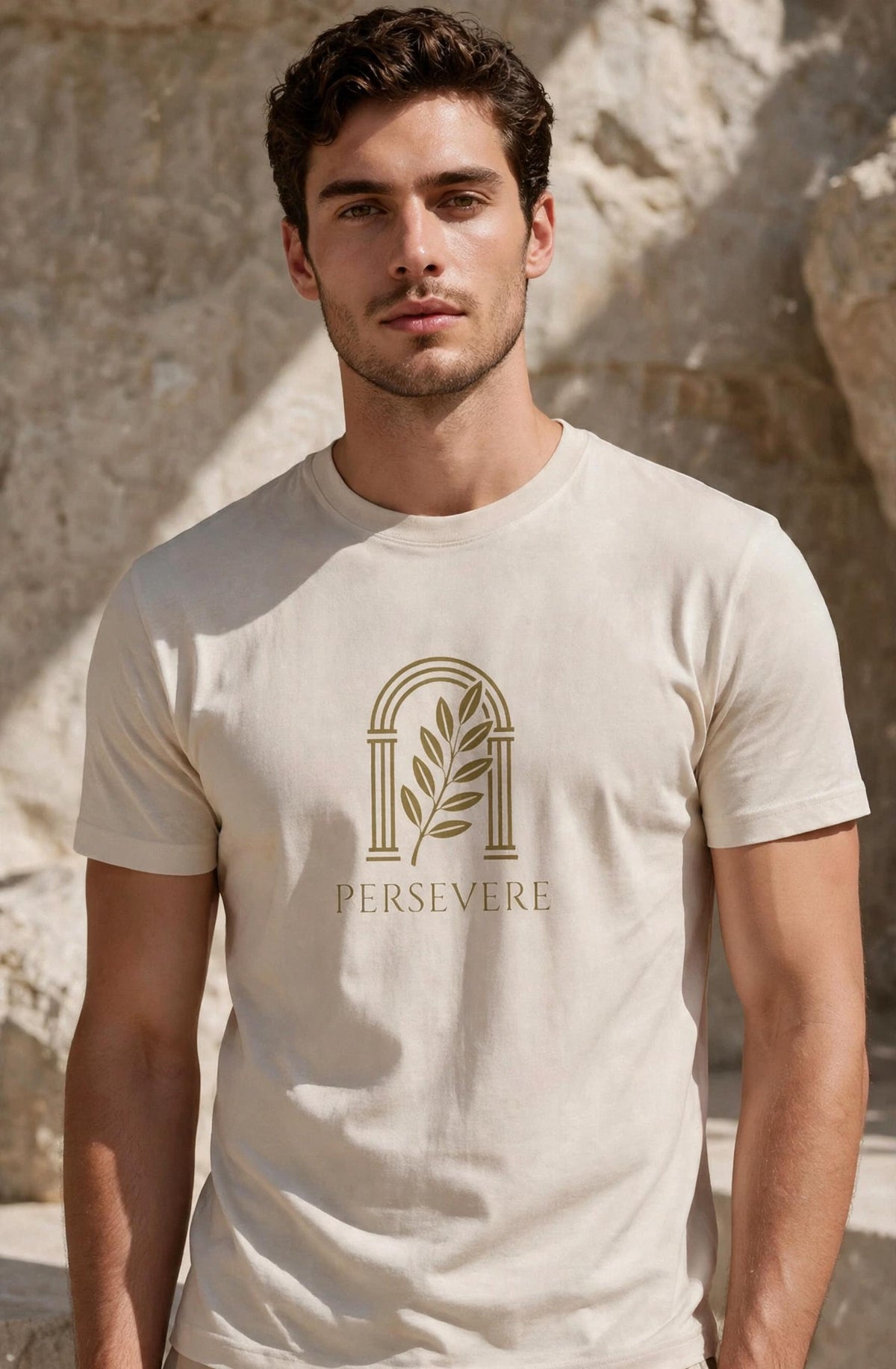 A man wearing a light beige crewneck t-shirt with a gold graphic of an arch and leaf and the word 'PERSEVERE' printed on the chest, set against a stone wall background.