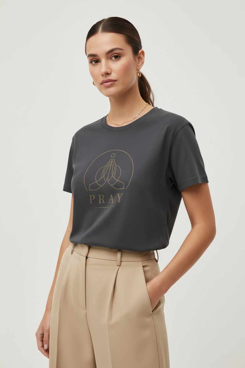A woman modeling a dark gray crewneck t-shirt with a gold graphic of praying hands and the word 'PRAY' on the front, paired with beige high-waisted trousers, against a light gray studio background.