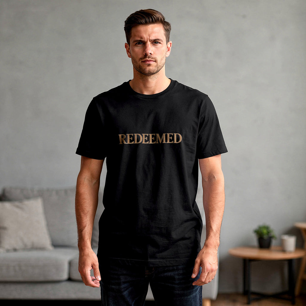 Man wearing a black t-shirt with 'REDEEMED' in gold letters, standing in a room with a gray wall and a couch.