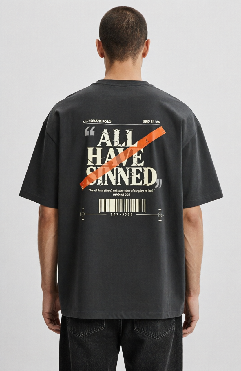 A man viewed from behind wearing a dark gray oversized graphic T-shirt with a large white text print reading 'ALL HAVE SINNED' crossed out in orange, paired with dark jeans against a light gray studio background.