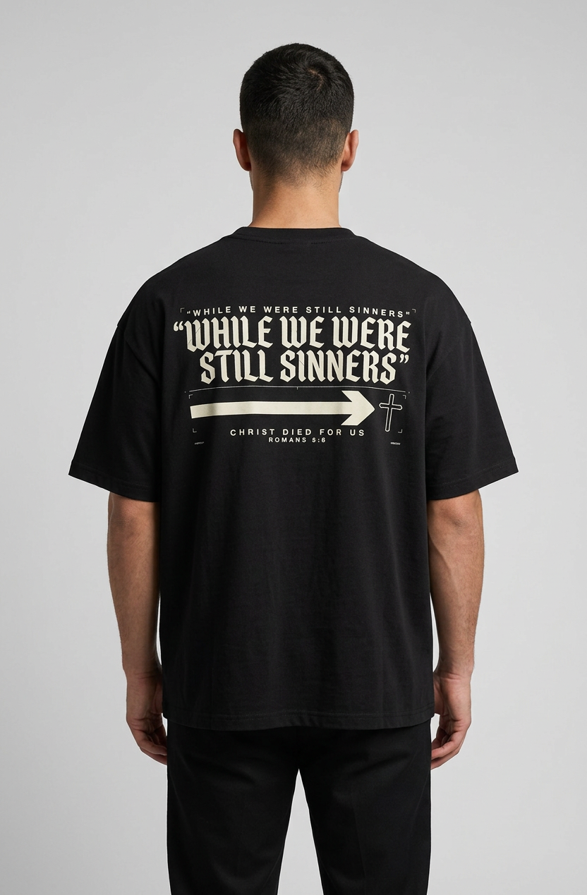A man viewed from behind wearing a black t-shirt with a large white graphic print on the back that reads 'WHILE WE WERE STILL SINNERS' and 'CHRIST DIED FOR US', paired with black pants against a light gray studio background.