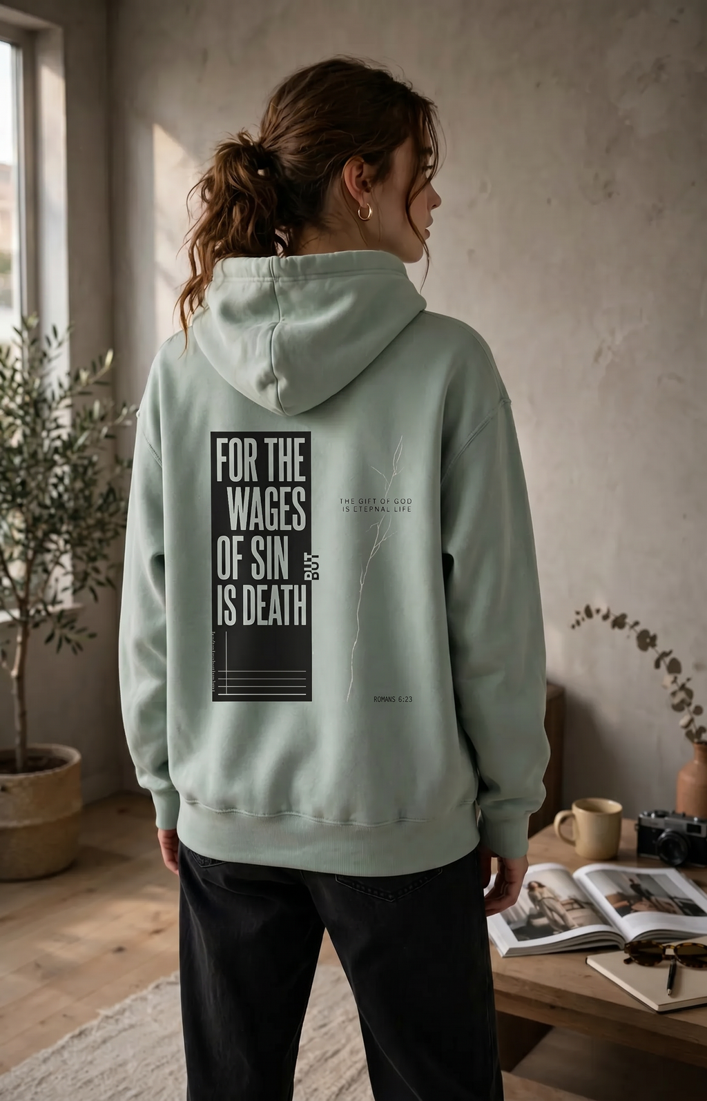 Person wearing a light green hoodie with text on the back in a room with a table and plant.