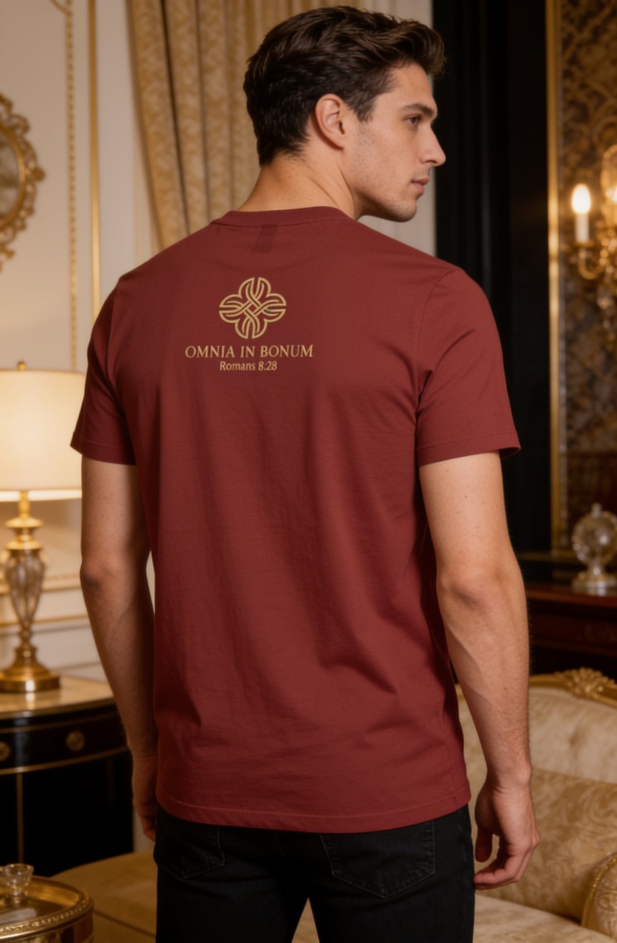 A man wearing a burgundy t-shirt with a gold Omnia in Bonum logo on the back, standing in an elegant, vintage-style room with gold curtains and lamps.
