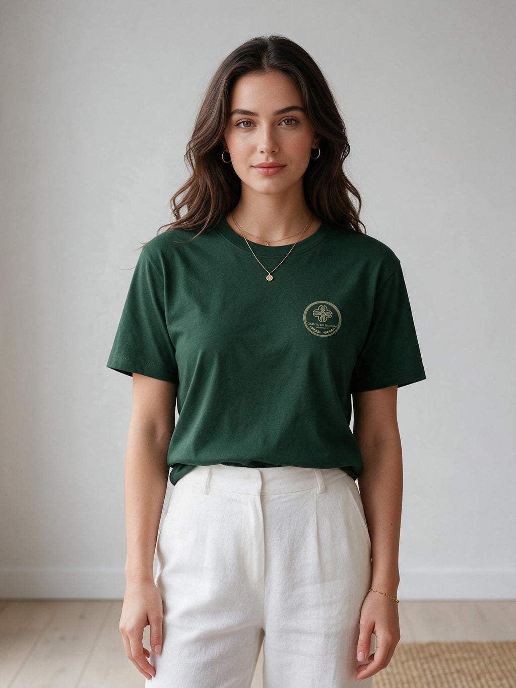 A woman wearing a dark green short-sleeve t-shirt with a small gold circular logo on the left chest, paired with white trousers, standing against a neutral light gray wall.