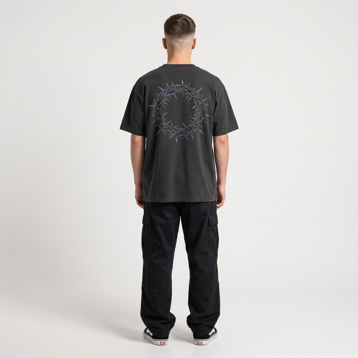 A man viewed from behind wearing a dark charcoal gray t-shirt with a purple crown of thorns graphic on the back, paired with black cargo pants and white sneakers, against a plain white studio background.