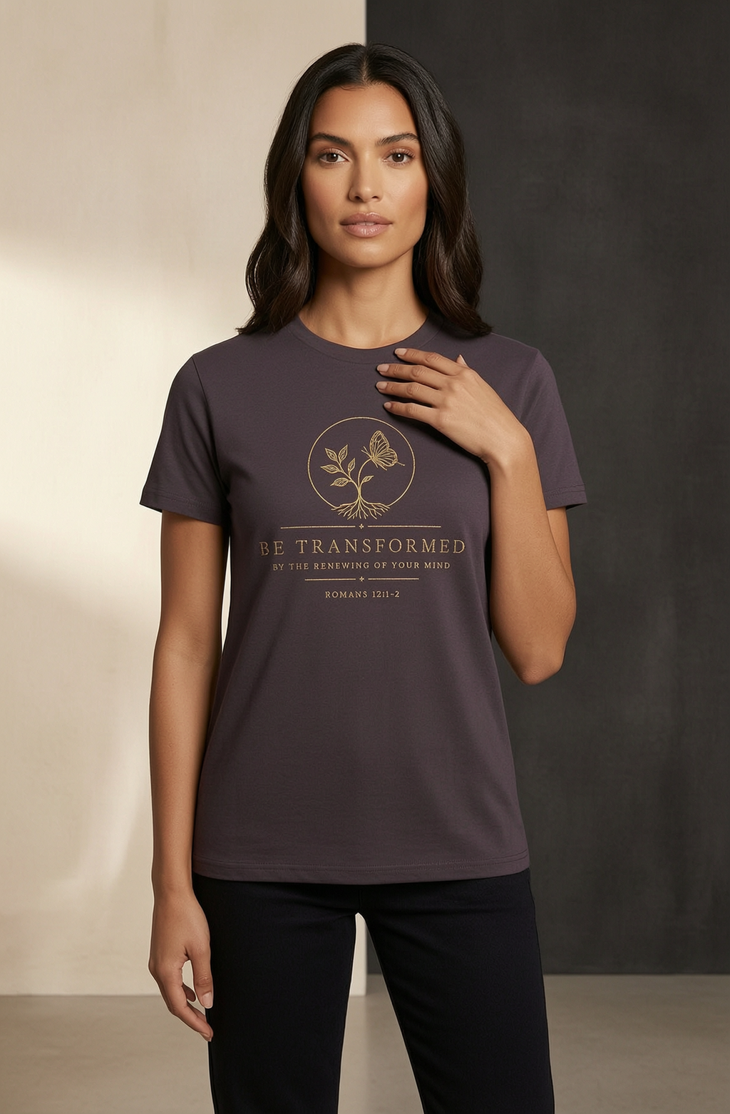A woman modeling a dark plum purple short-sleeve t-shirt with a gold butterfly graphic and the text 'BE TRANSFORMED' on the front, paired with black pants against a split beige and dark gray studio background.