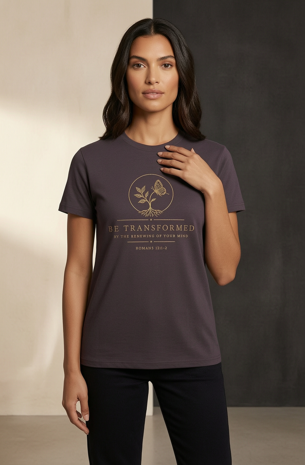 A woman modeling a dark plum purple short-sleeve t-shirt with a gold butterfly graphic and the text 'BE TRANSFORMED' on the front, paired with black pants against a split beige and dark gray studio background.