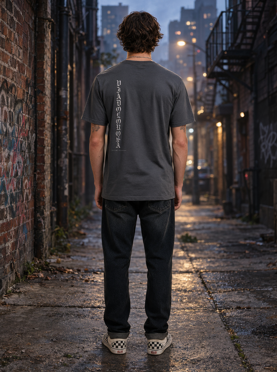 A man viewed from behind wearing a dark gray t-shirt with vertical white text on the back, dark jeans, and checkered sneakers, standing in a wet urban alleyway at dusk.