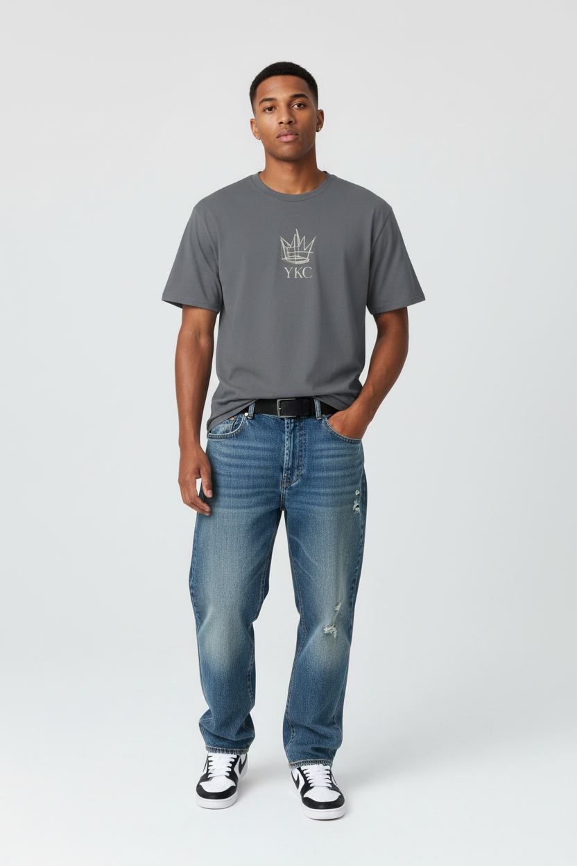A man modeling a dark gray crewneck t-shirt with a white crown and 'YMC' logo on the chest, paired with blue jeans and black-and-white sneakers, against a light gray studio background.