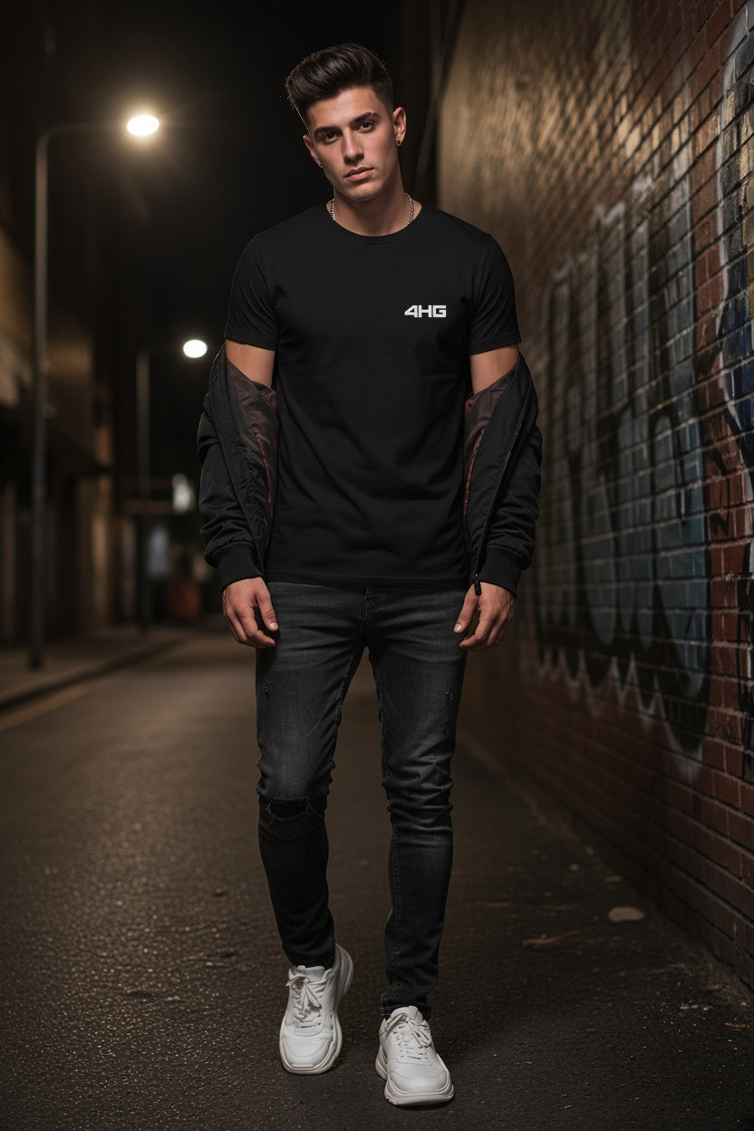 A man wearing a black crewneck t-shirt with a small white '4HG' logo on the chest, styled with a black long-sleeve shirt draped off his arms, black jeans, and white sneakers, walking down a dark brick alleyway at night.