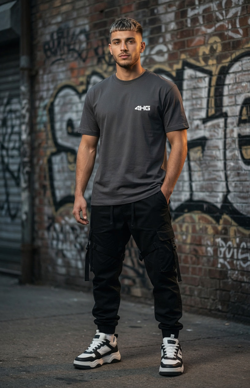 A man wearing a dark gray crewneck t-shirt with a white '4HG' logo on the chest, paired with black cargo pants and white sneakers, standing in front of a graffiti-covered brick wall.