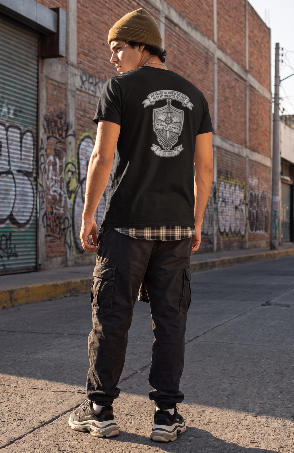 A man standing on an urban street, viewed from behind, wearing a black t-shirt with a large white graphic print on the back, black cargo pants, and a brown beanie.