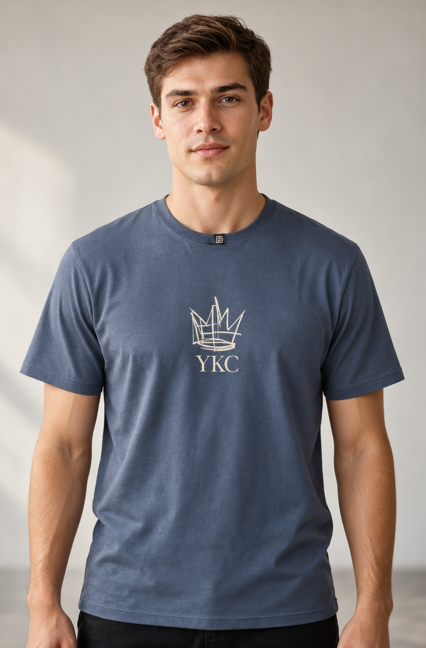 A man wearing a slate blue crewneck t-shirt with a white printed crown logo and the letters 'YKC' on the chest, photographed against a neutral studio background.